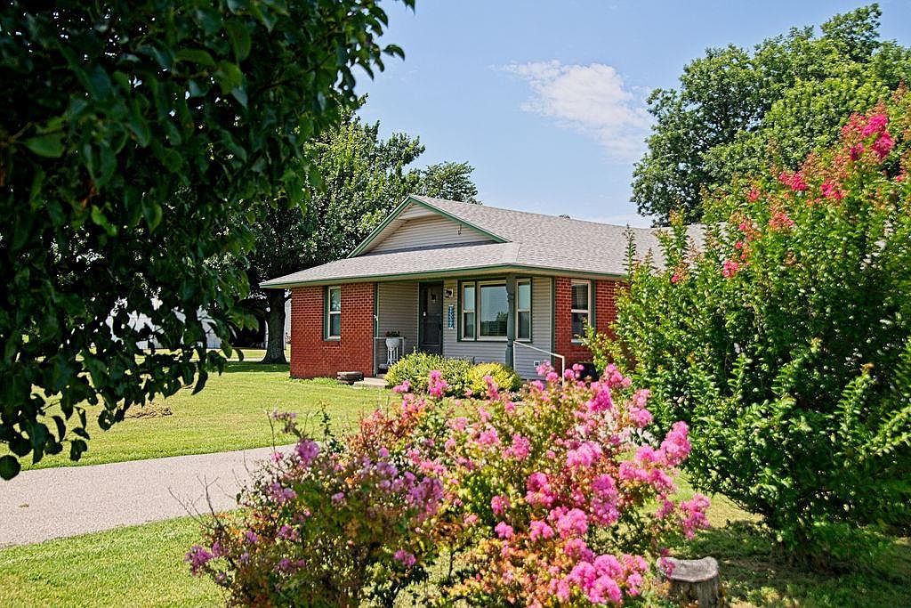 The 1960 brick farmhouse at 6001 N Midwest Blvd framed by mature crepe myrtles in full pink bloom